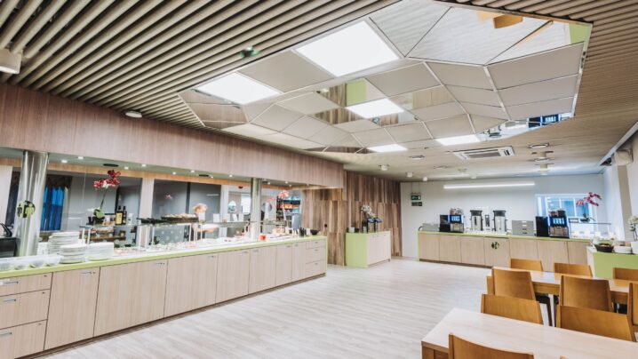 Moderni ruokasali, jossa on puukalusteet, geometrinen kattosuunnittelu, tarjoilutasot ja kahvinkeittimet.
