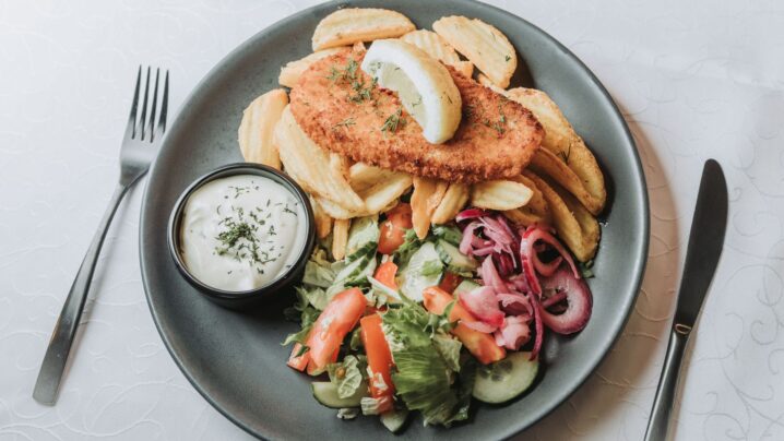 Paneeritud kala, mida serveeritakse friikartulite, salati ja väikese kausitäie kastmega tumehallil taldrikul kahvli ja noaga valgel laudlinal.