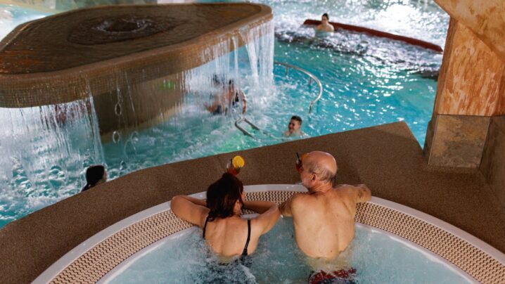 An old couple sitting with their backs in the jacuzzi above and looking down at what is happening in the pool.