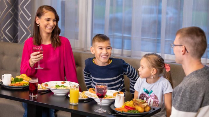 Perhe, joka nauttii ateriasta ruokapöydän ääressä, pöydällä erilaisia ​​ruokia ja lasit punaisia ​​ja oransseja juomia. Taustalla moderni harmaa sisustus.