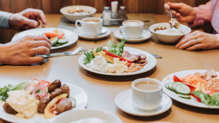 Ihmiset nauttivat aamiaisesta. Lautasilta näkyy munat, pekoni, pavut ja salaatti sekä kupit kahvia.