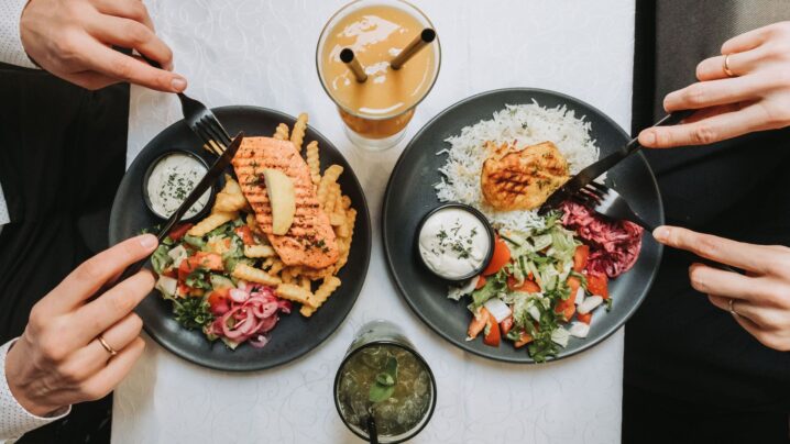 Kaksi lautasta täynnä ruokaa. Toinen ruokalaji perustuu riisiin ja toinen ranskalaisiin perunoihin. Ruoka on tummanharmaalla lautasella. Lautasten ääressä on myös kaksi värikästä cocktailia.